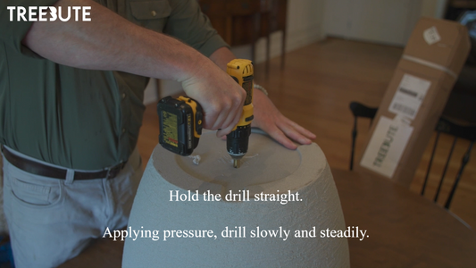 Man drilling holes with a Dewalt power tool indoors at a wooden table. Treebute Kit package rests on a chair in the background as a meaningful memorial tree gift option. 