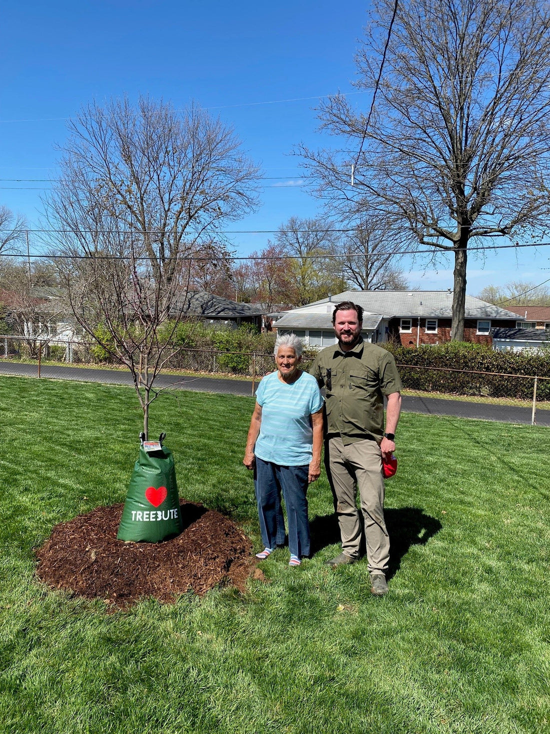 Elderlywoman and young man stand next to a newly planted Eastern Redbud Treebute memorial tree in a residential yard on a sunny day with neighborhood houses and street in the background