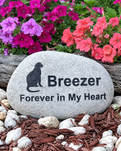 A custom engraved memorial garden stone featuring a black silhouette of a dog and the text 'Breezer Forever in My Heart', displayed in a garden bed with mulch, white stones, and vibrant pink and red flowers.