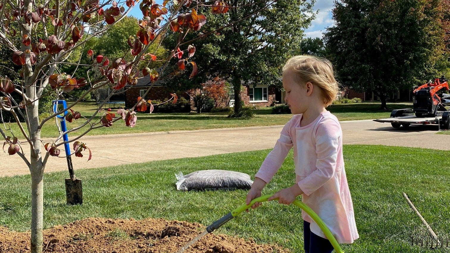 Child watering a newly planted tree with a garden hose on a sunny day.
